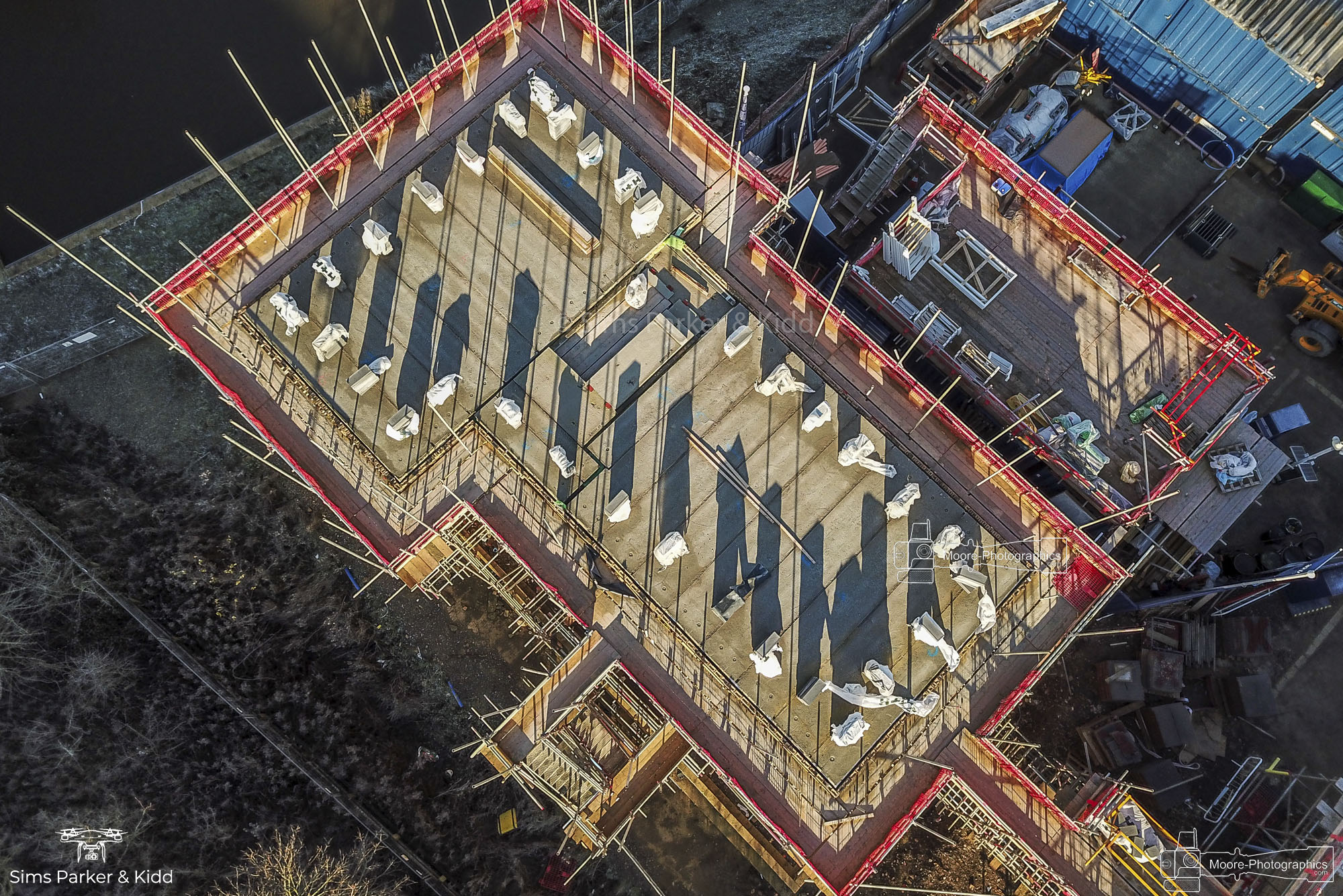 Overhead drone view of a timber-framed building under construction, red edge protection and scaffolding around the open floor plates